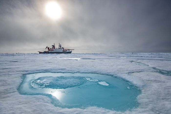 The Polarstern is visible on the horizon beyond a semi-frozen melt pond. Photo courtesy Lianna Nixon, 2020 (CIRES and AWI).