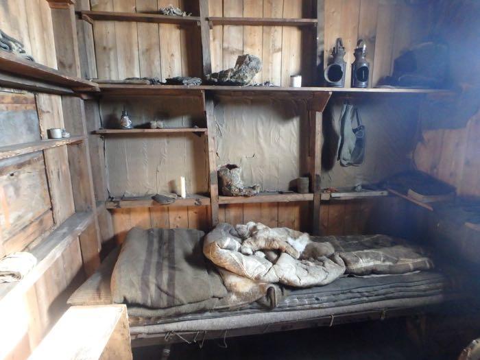 A bed and some wooden shelves in an old expedition hut.