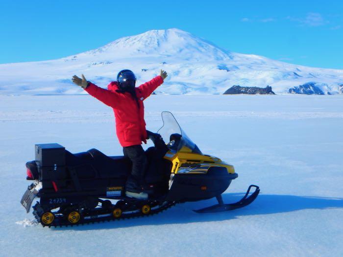 Person with arms raised on a snow machine on the sea ice. A volcanic mountain is in the background.