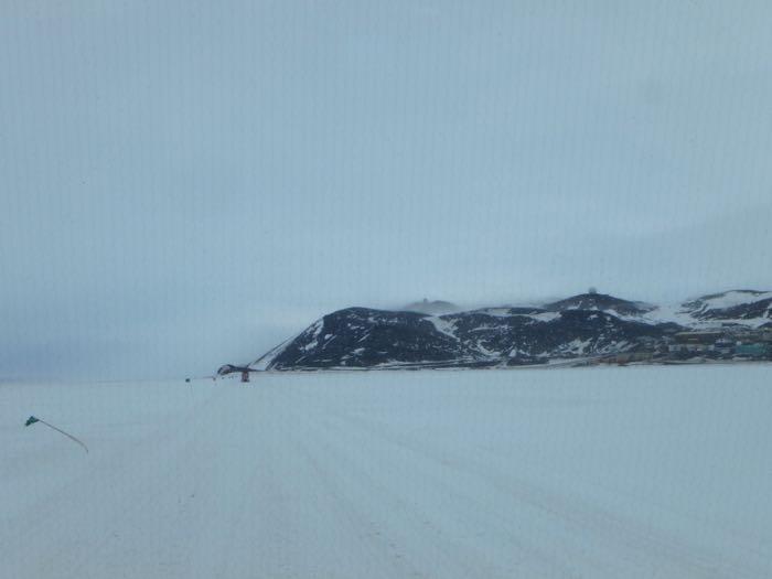 Pisten Bully driving towards McMurdo station. Flag bent over due to wind and gray skies.