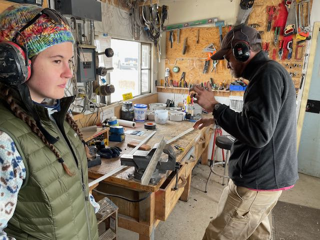 Sarah and Jeremy stand in a carpenter's workshop. Sarah stands to the left and looks off into the distance, while Jeremy is on the right and is looking at an object in his hand. 
