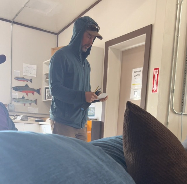 A man with a blue hoodie and a baseball cap stand holding a walkie talkie and a piece of paper. 