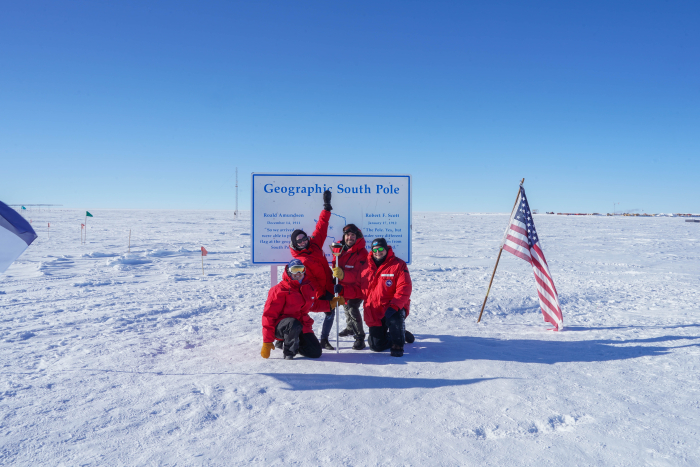 A group of people stand in front of the geographic South Pole