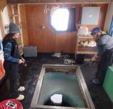 Amy Osborne and Denise Hardoy standing inside a dive hut fishing in an icy dive hole.